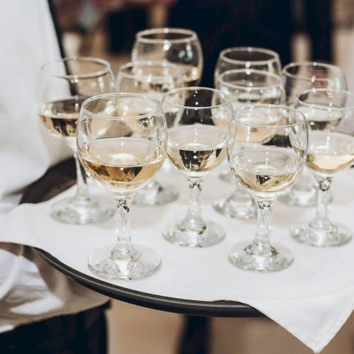 A waiter carrying a black tray with several white wine glasses on a white napkin, filled with pale golden wine, at a formal event.