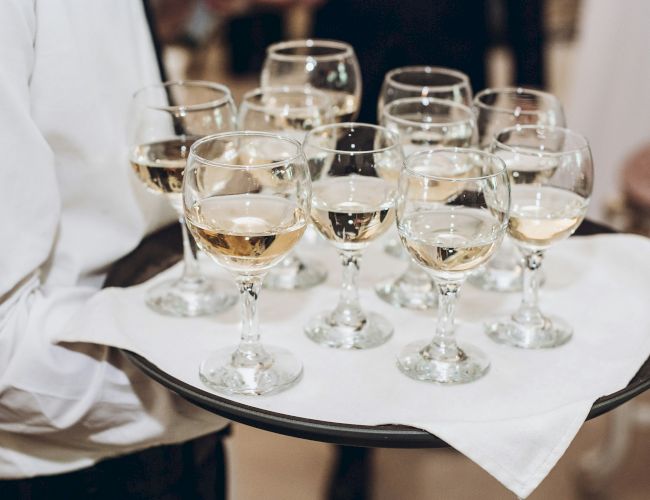 A waiter carrying a black tray with several white wine glasses on a white napkin, filled with pale golden wine, at a formal event.