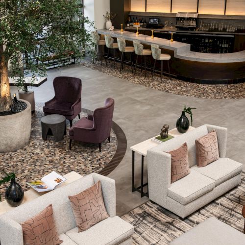 A luxurious hotel lounge with a curved bar, high stools, plush sofas, earthy tones, potted trees, and warm lighting.