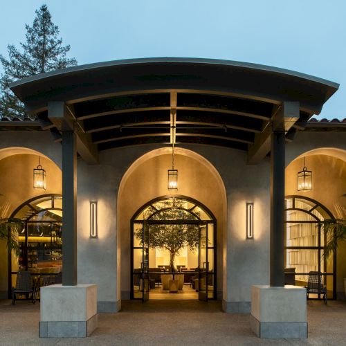 A stylish, arched entrance to a modern building with warm lighting and indoor greenery, under a curved roof, inviting interior visible.