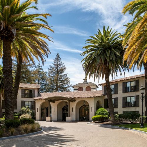 A sunny hotel courtyard with palm trees, a circular driveway, and a central arched entrance.