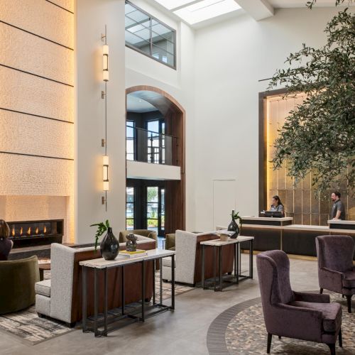 A modern hotel lobby with tall windows, a large tree indoors, stylish seating, and a reception desk in the background, people lounging and walking.
