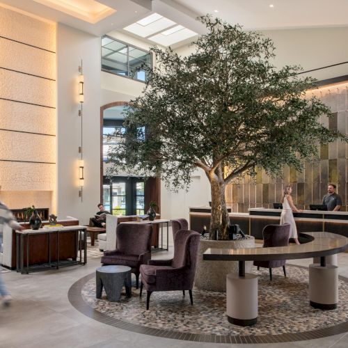 A luxurious hotel lobby with a large potted tree centerpiece, modern seating around a circular rug, a receptionist desk, and guests mingling near marble walls and warm lighting.
