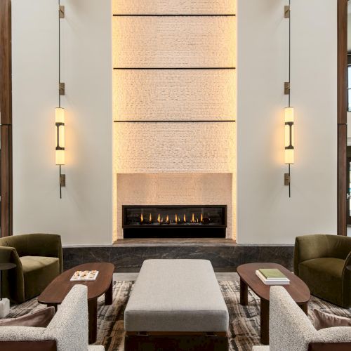 A modern hotel lobby with a central glowing fireplace, symmetrical seating, and tall vertical light fixtures flanking a white feature wall.