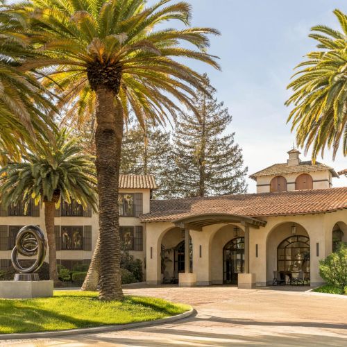 A sunny campus with palm trees, a circular driveway, and a beige building with arched entrances; a statue sits on the left.