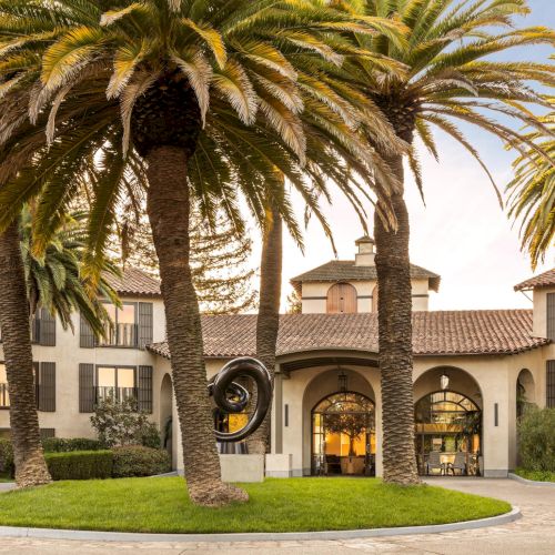 A hotel-like building with palm trees lining the driveway and a circular grassy island in front, arched entrance, sunny day.