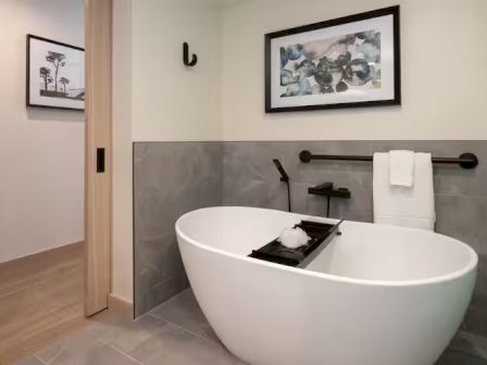 A modern bathroom with a freestanding white oval tub, wall-mounted faucet, gray tile wainscoting, towel rack, and framed art above, plus a doorway to another room.