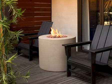 Two black wooden chairs sit around a round concrete fire pit with flames, on a shaded patio next to a wooden wall and some greenery.