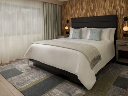 A modern hotel bedroom with a large white bed, padded headboard, two nightstands, warm lamps, and a textured accent wall by the window.