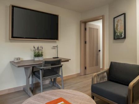 A small modern living area with a wall-mounted TV, a desk and chair, a gray armchair, a round coffee table, and a doorway leading to another room.