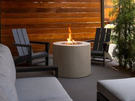 A modern outdoor seating area with a round fire pit, gray cushioned sofas, wooden privacy wall, and two dark chairs around the flame, cozy evening vibes.