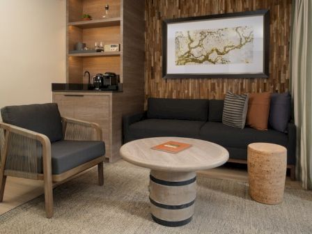 A modern living room with a black sofa, wooden armchair, round stone coffee table, a textured wood accent wall, and a small cork stool, neatly arranged.