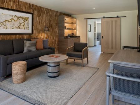 A modern living room with a dark sofa, a round coffee table, a textured wood accent wall, a beige rug, and a small armchair; a dining area and sliding barn door in the background.