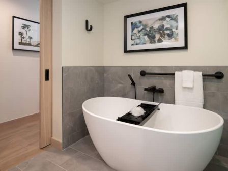 A modern bathroom with a freestanding white oval tub, wall-mounted faucet, towel bar, framed art, and a small tray with toiletries over the tub.