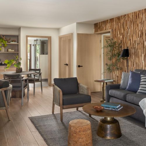 A stylish, airy living room with a gray sofa, wooden coffee table, woven rug, and a textured accent wall, plus a dining nook and shelves in the background, all bathed in natural light.