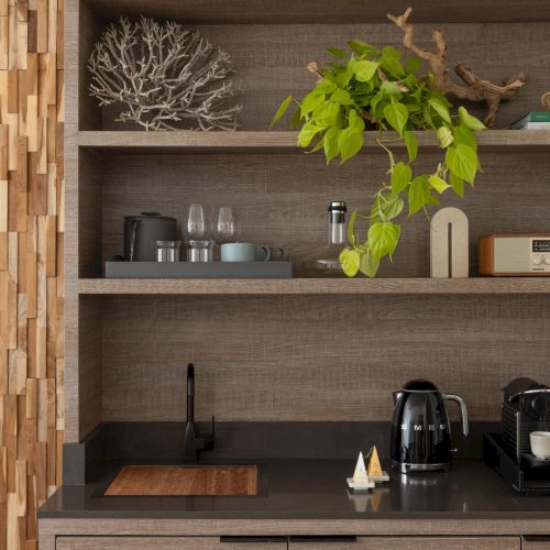A modern kitchen nook with wooden shelves, small plants, a kettle, toaster, cups, and decorative items on a dark countertop.