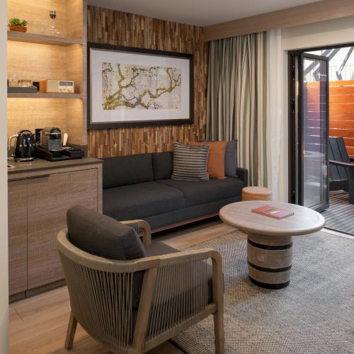 A modern hotel suite with a seating area, wood accents, and a small kitchenette; a round coffee table, sofa, armchairs, and a balcony with a fire pit.