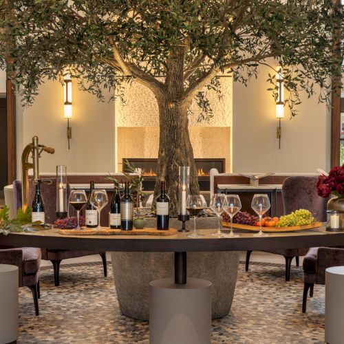 A group of people sits around a long dining table in a bright, airy room with large windows and plants; warm, casual gathering near sunset.