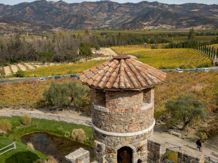 A stone fortress tower atop walls overlooking a vineyard and rolling hills under a blue sky, with visitors exploring the structure.