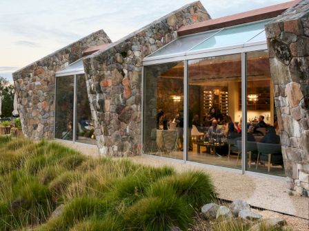 A stone, modern-looking building with large glass windows, overlooking grassy landscaping and a calm outdoor setting.